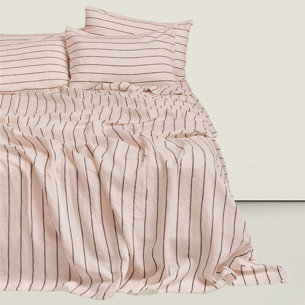 A bed dressed in a light blush pink striped bedding set, featuring a gathered ruched duvet cover and matching pillowcases, set against a solid light beige background.