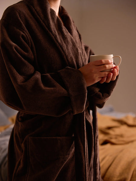 A person wearing a plush dark brown bathrobe and holding a white ceramic mug in a softly lit bedroom setting.